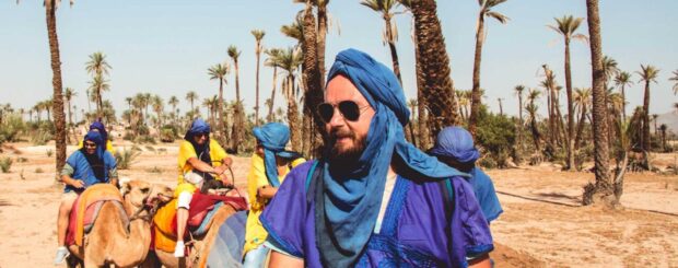 A group of camels with colorful blankets on their backs walking in a line on a sandy path surrounded by palm trees. In the background, a blue sky with white clouds can be seen. People are seated on top of the camels, enjoying the ride while wearing traditional clothing and headscarves. The scene suggests a peaceful and exotic experience in the Moroccan desert.