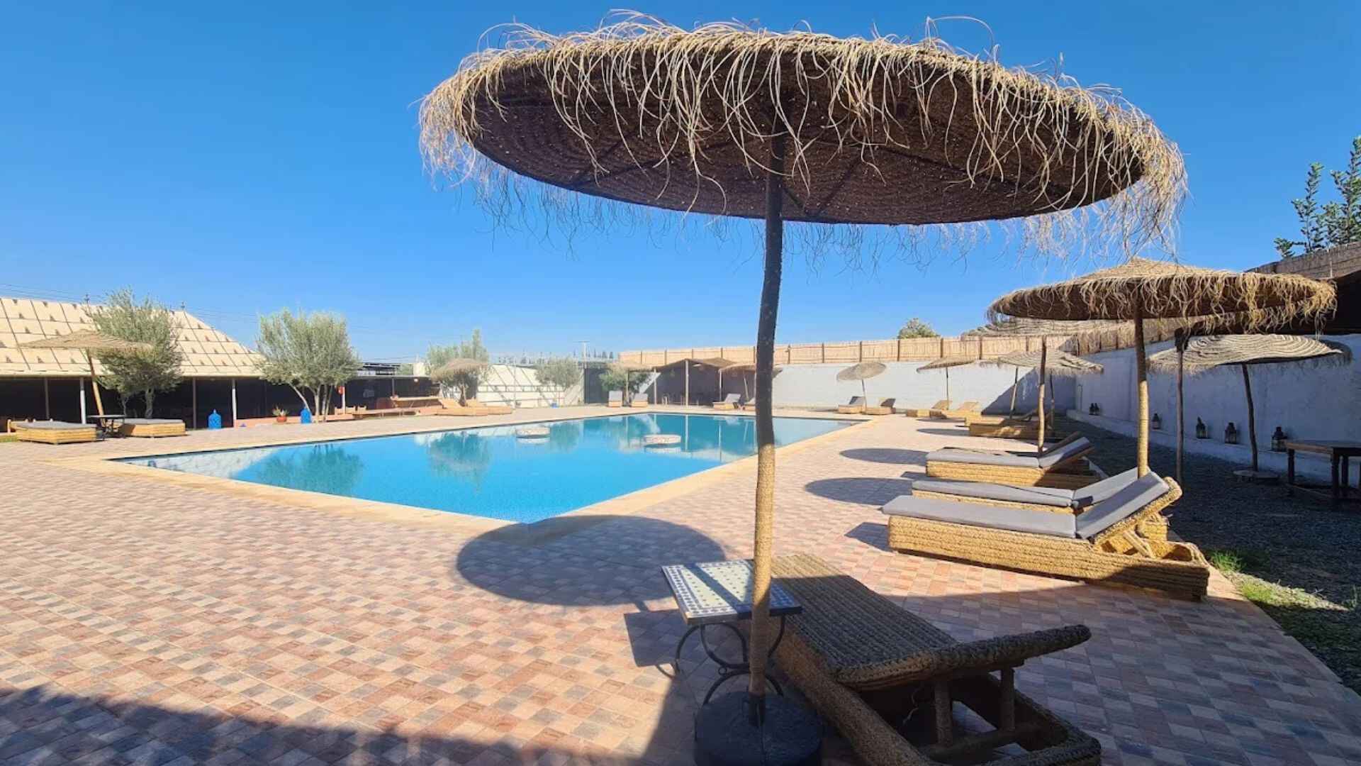 Outdoor swimming pool at Agafay Desert camp with sun loungers and straw parasols under clear blue sky near Marrakech.